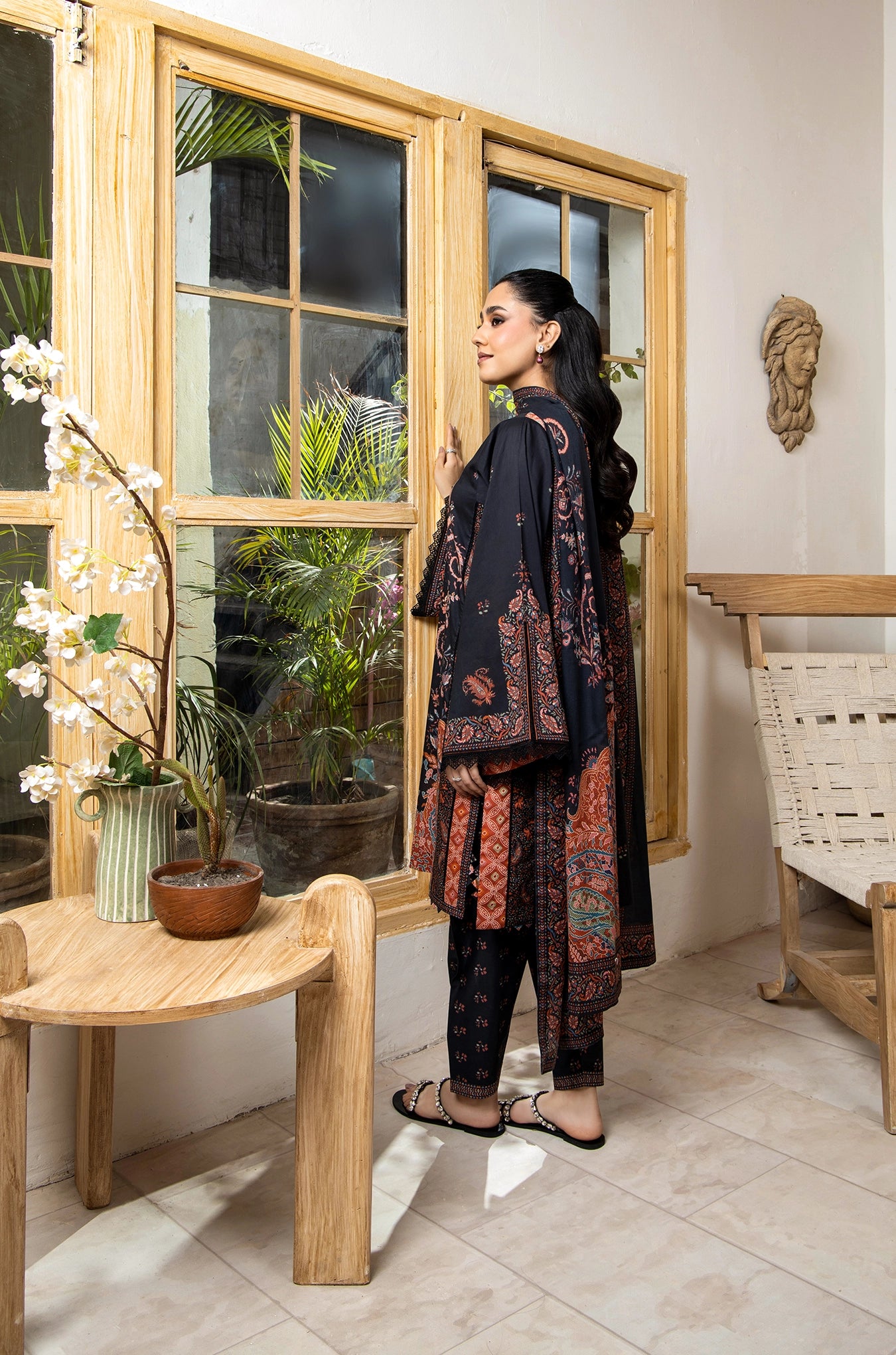 Woman in a patterned outfit standing in a room with wooden furniture and plants.