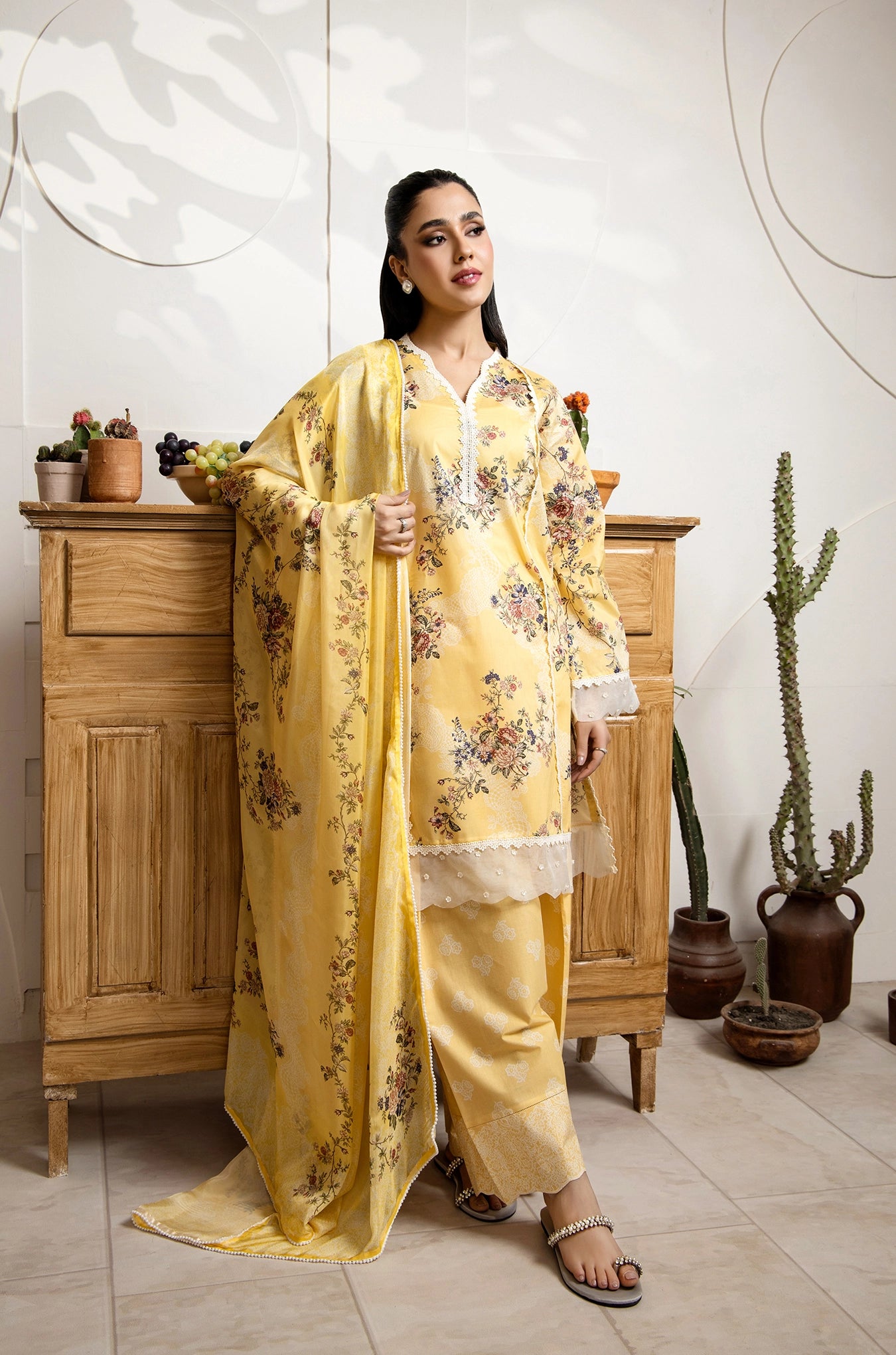 Woman in a yellow floral outfit standing in a room with wooden furniture and plants.