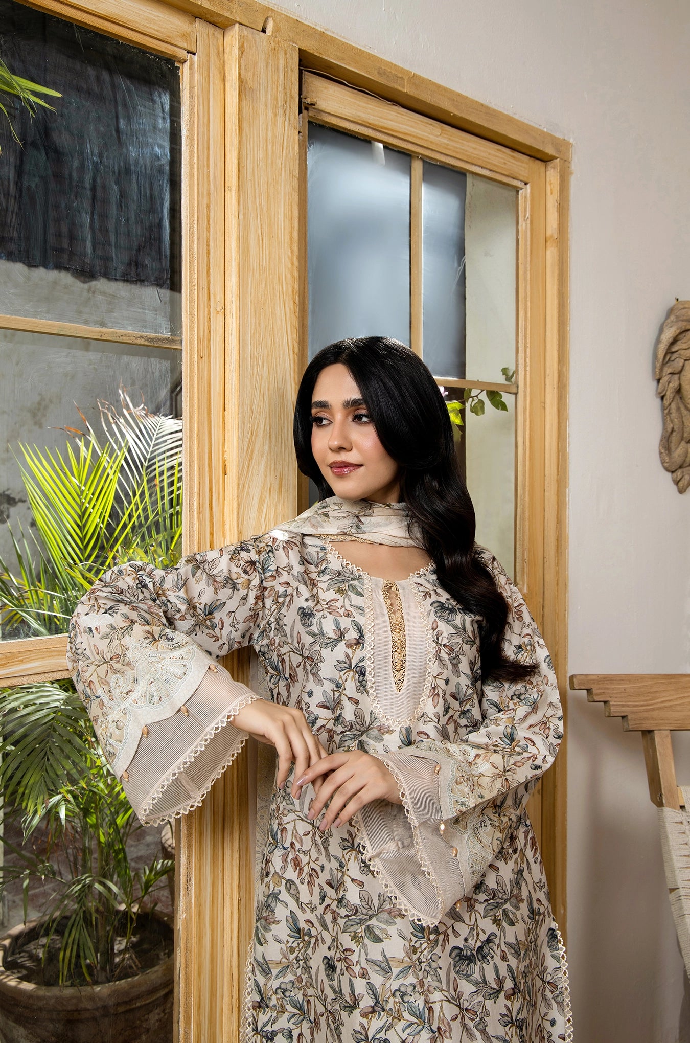 Woman in a brown patterned dress standing indoors with plants and a window in the background