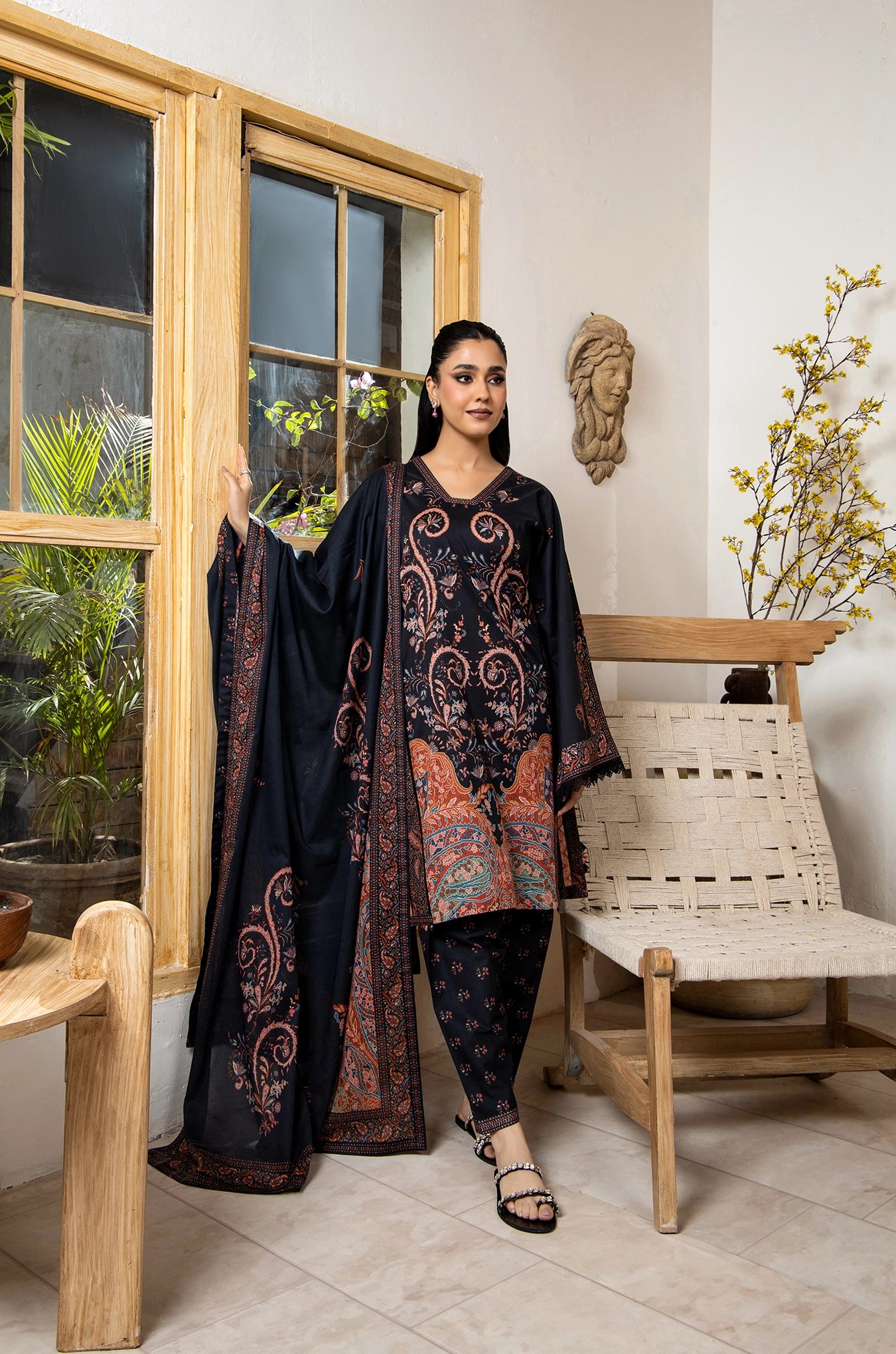 Woman in traditional attire standing in a room with wooden furniture and plants.