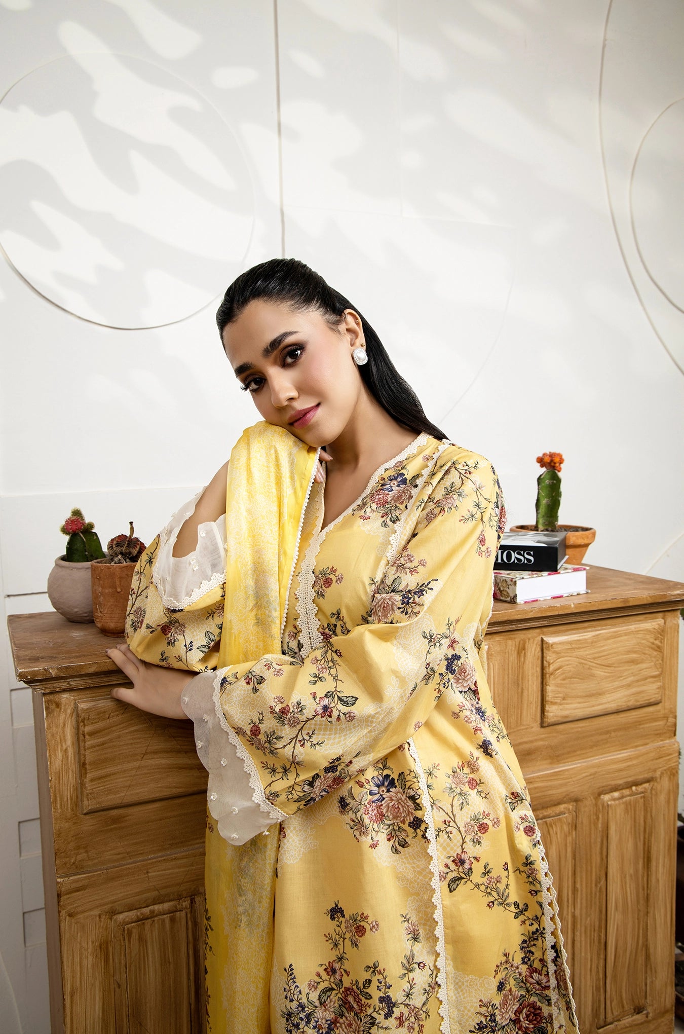 Woman in a yellow floral dress standing in a room with wooden furniture and decorative items.
