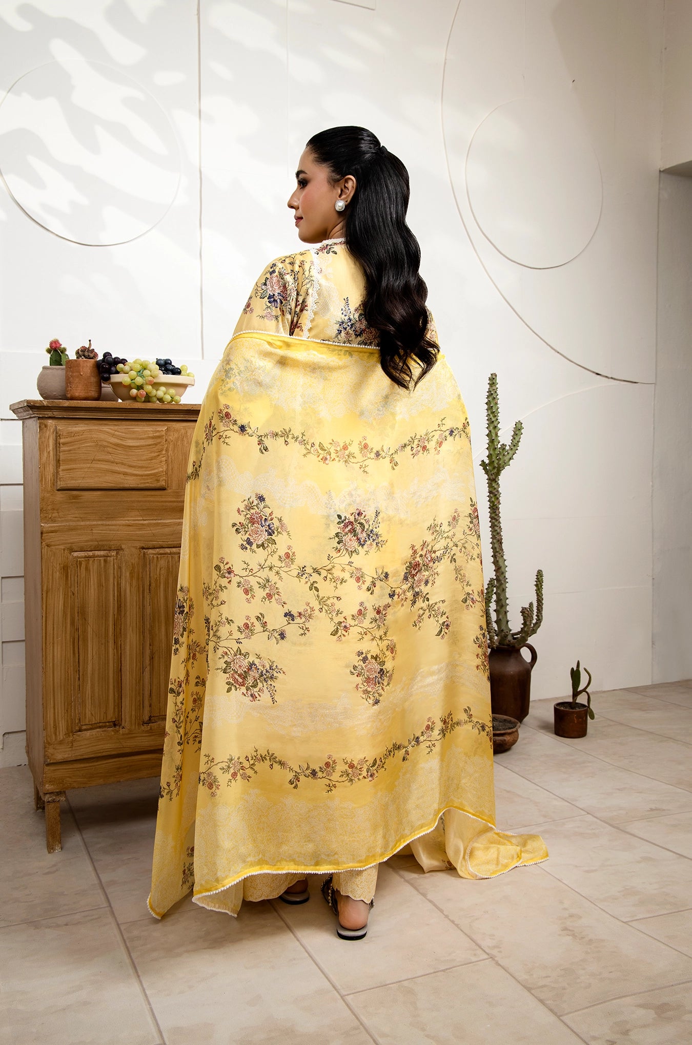 Woman in a yellow floral dress standing in a room with wooden furniture and plants.
