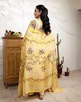 Woman in a yellow floral dress standing in a room with wooden furniture and plants.